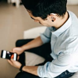 Younger man wearing a hearing aid while looking at his phone and sitting on the edge of a bed